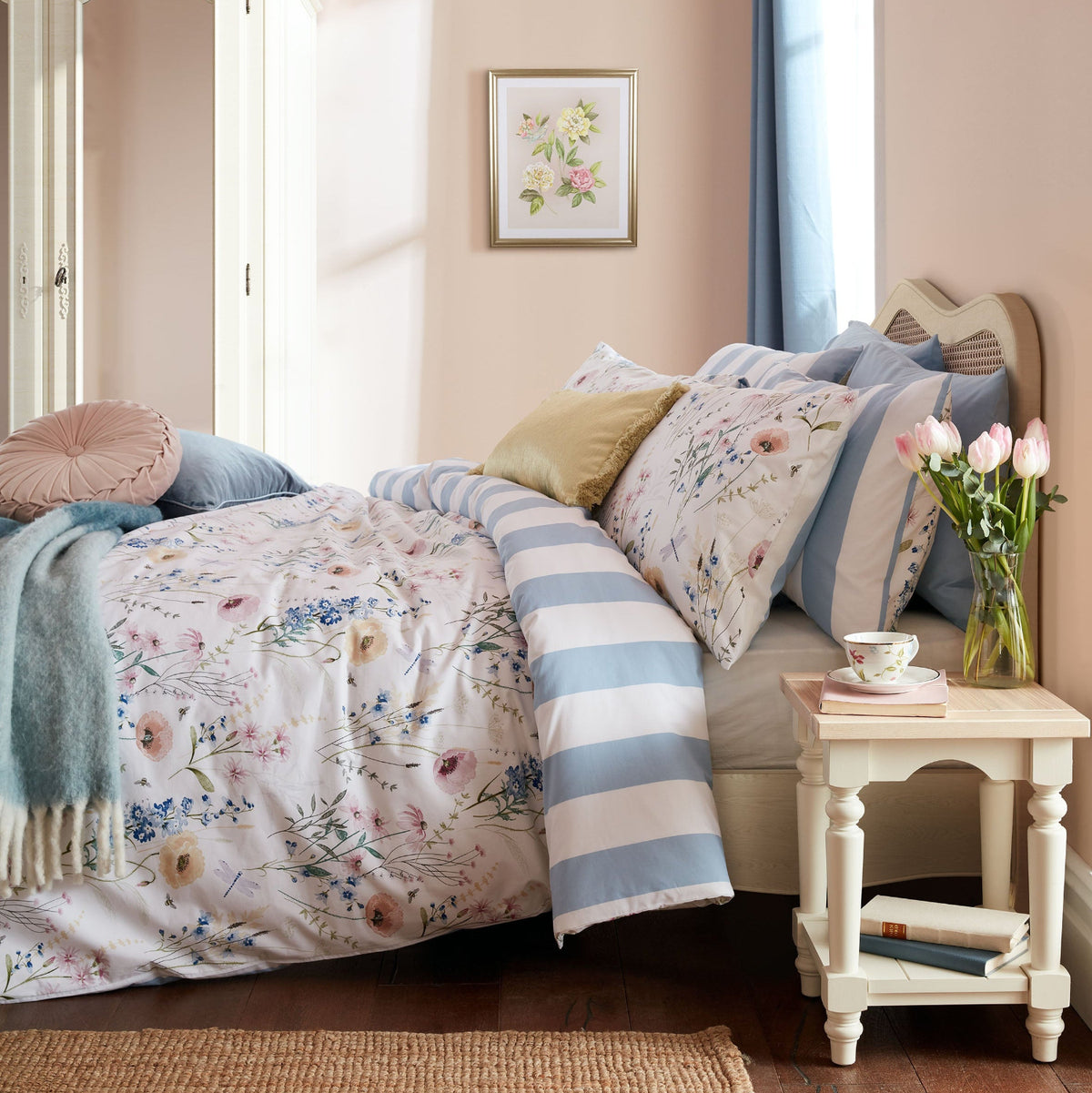 Bedroom set with floral and blue striped bedding, pillows, and a bedside table with tulips.