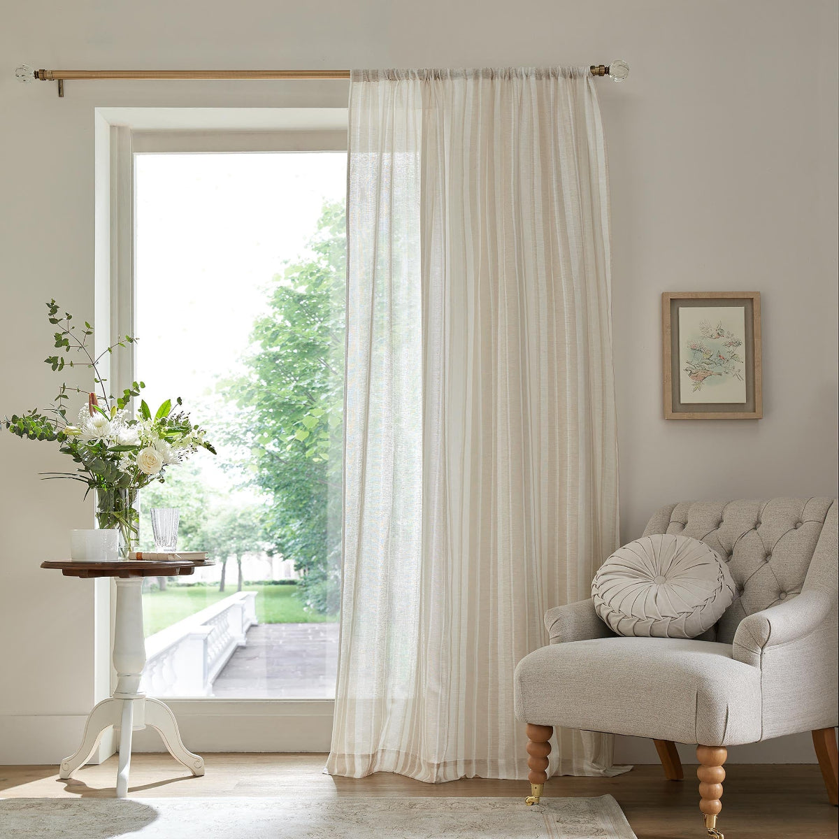 Sheer cream striped curtains hanging by a window next to a tufted armchair and side table.